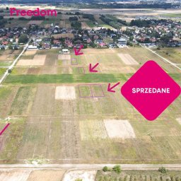 Marcin Kucharczyk Freedom Nieruchomości - Zdjęcie z lotu ptaka na pola z zaznaczonymi działkami na sprzedaż i napisem 'Sprzedane' w różowym rombie, z widocznymi domami w tle.