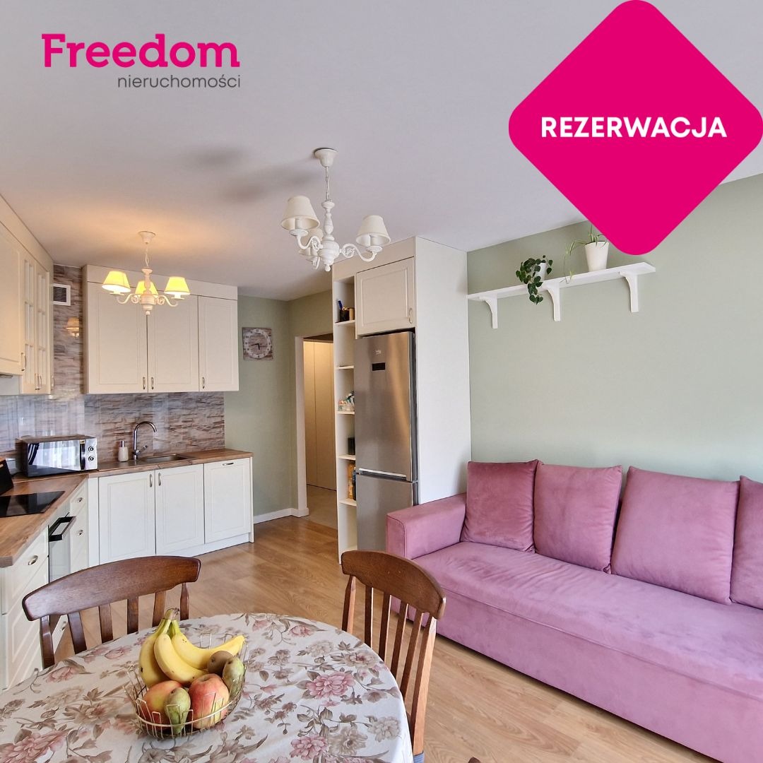 Jasne wnętrze: kuchnia z jadalnią i różową sofą. Na ścianie napis 'Rezerwacja'. Na stole owoce w koszyku. Widok na umeblowane mieszkanie z oferty Freedom nieruchomości.