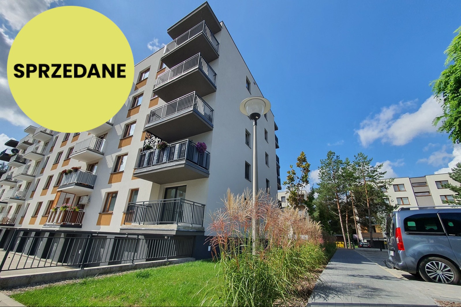 Nowoczesny budynek wielorodzinny z balkonami, oznaczony okrągłą naklejką 'Sprzedane' na elewacji. Widok z dołu, błękitne niebo w tle.