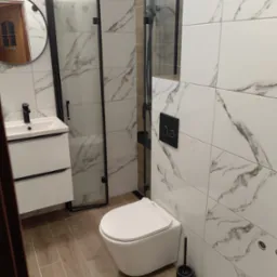 Nowoczesna łazienka z kabiną prysznicową typu walk-in, białą armaturą i płytkami imitującymi marmur. Czarna rama kabiny i dodatki, drewnopodobna podłoga.