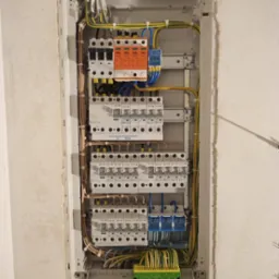 Szczegółowe ujęcie na otwartą skrzynkę elektryczną z widocznymi bezpiecznikami, przewodami i elementami montażowymi wewnątrz wnęki ściennej.