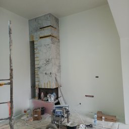 PPK PERFEKT 22 - Obudowa kominka z marmurowych płyt w trakcie montażu, zabezpieczona taśmą. W tle ściana przygotowana do malowania, widoczne narzędzia i akcesoria malarskie.