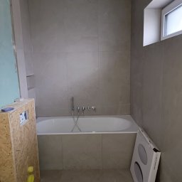 PPK PERFEKT 22 - Wykończona łazienka z wanną w zabudowie, płytki w odcieniach beżu, okno. Częściowo widoczna obudowa instalacji i karton z lampami. Ujęcie z góry, neutralne oświetlenie.