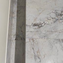 PPK PERFEKT 22 - Ściana wyłożona białymi płytkami imitującymi marmur z wnęką i czarnymi elementami dekoracyjnymi. Widoczne żółte taśmy zabezpieczające.