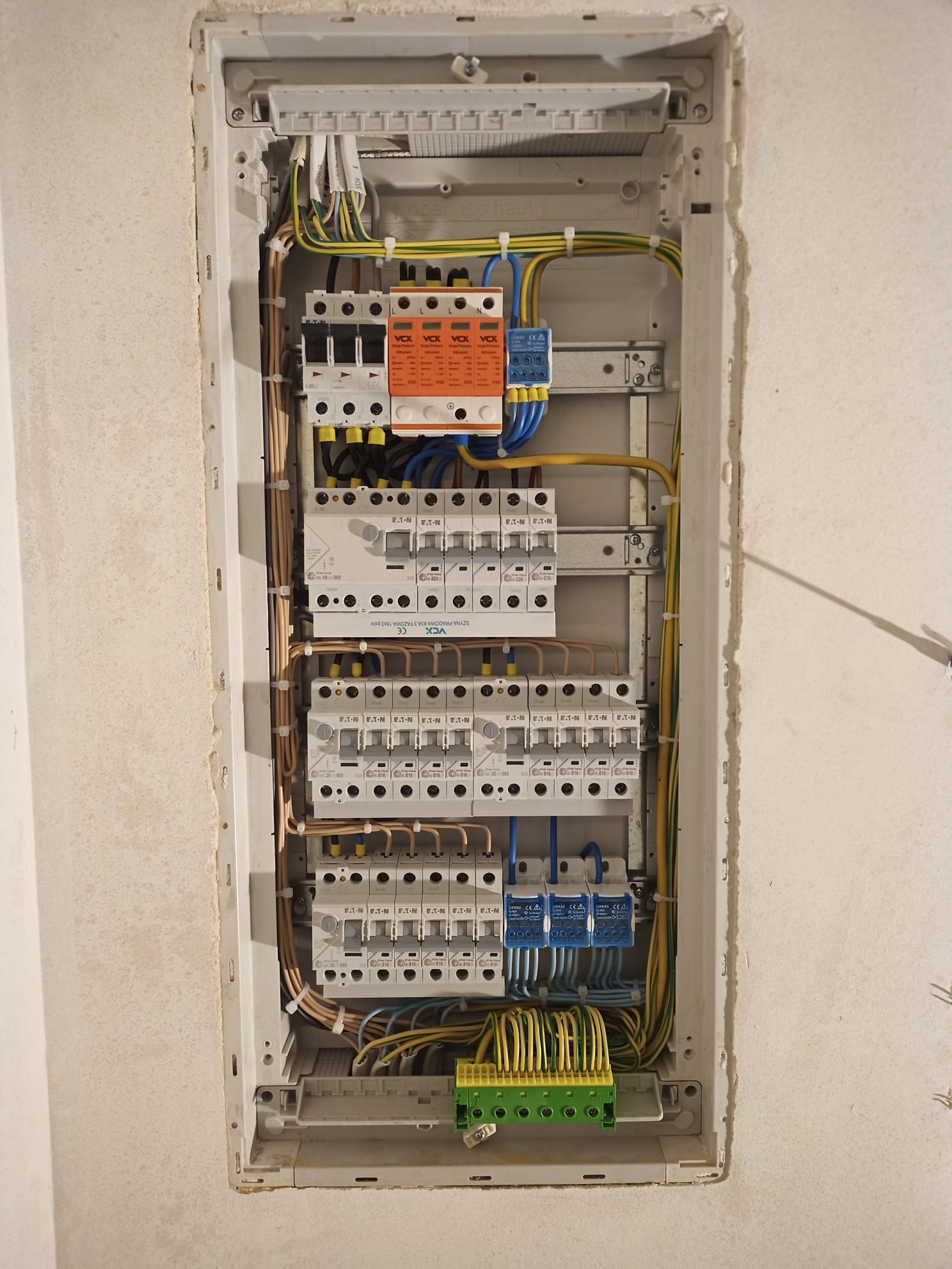 Szczegółowe ujęcie na otwartą skrzynkę elektryczną z widocznymi bezpiecznikami, przewodami i elementami montażowymi wewnątrz wnęki ściennej.