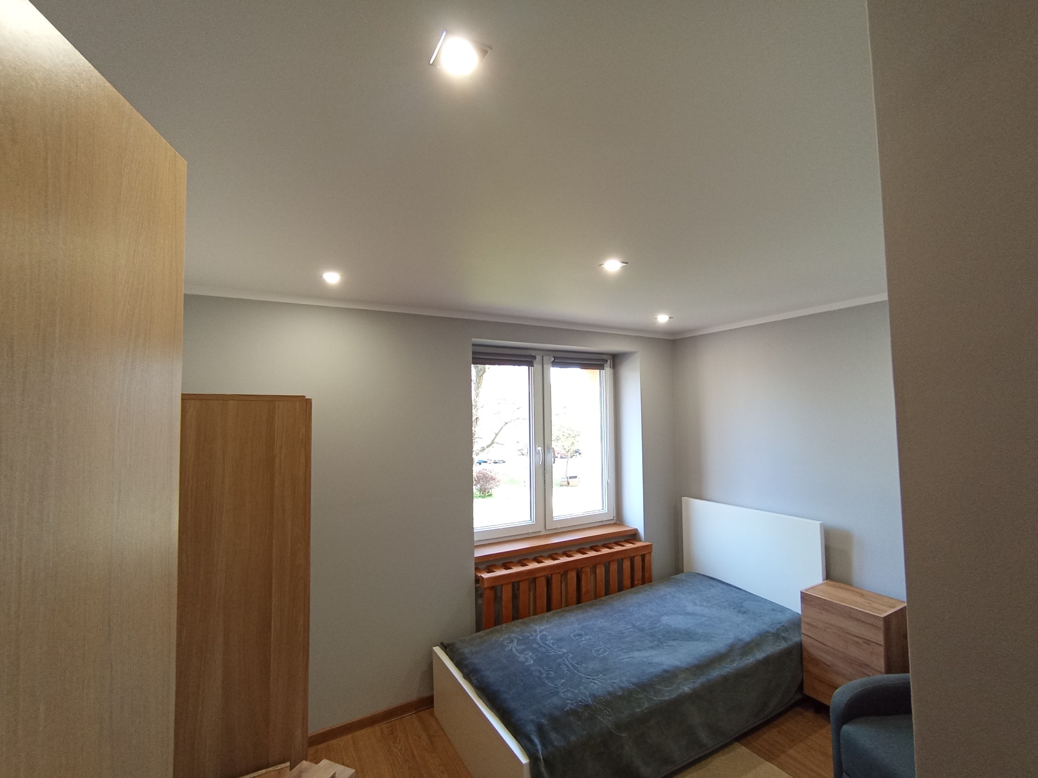 Pokój po remoncie z drewnianą szafą, łóżkiem z szarą narzutą i drewnianym grzejnikiem pod oknem. Ściany pomalowane na szaro, oświetlenie sufitowe punktowe.