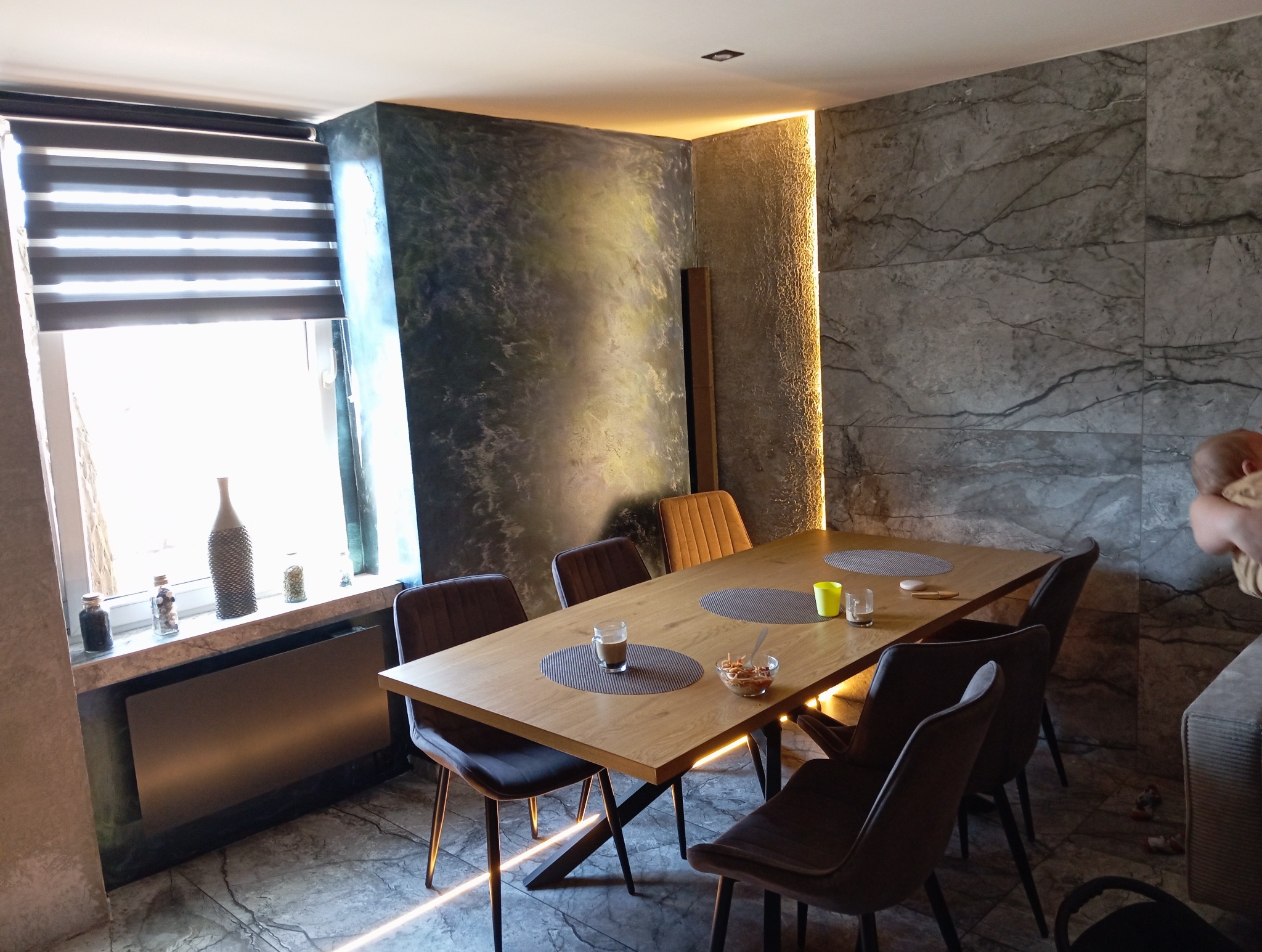 Jadalnia z drewnianym stołem, krzesłami i dekoracyjną ścianą z efektem marmuru. Okno z żaluzjami i dekoracje na parapecie. Oświetlenie punktowe.