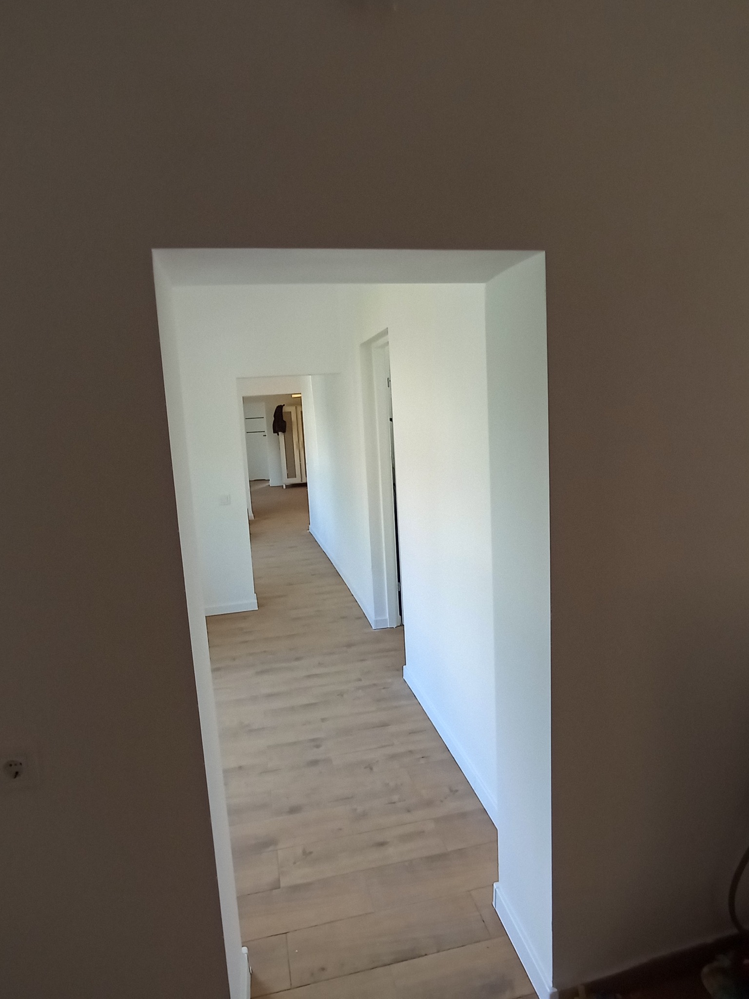 Wnętrze odnowionego mieszkania z drewnianą podłogą i świeżo pomalowanymi na biało ścianami, widoczne przez przejście z brązową ścianą po lewej.