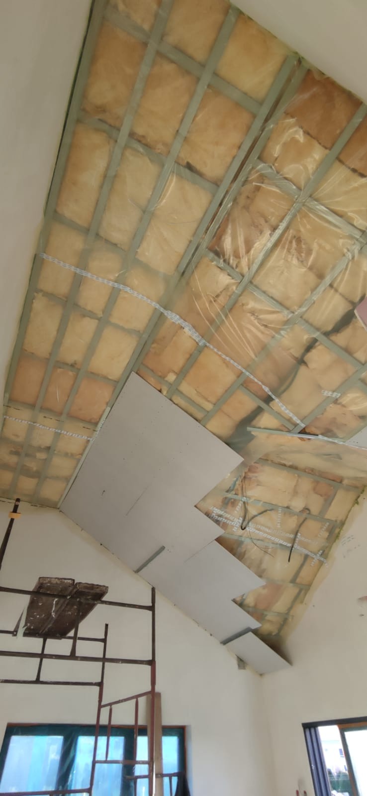 Skośny sufit z izolacją i częściowo zamontowaną płytą karton-gips, widok z dołu, rusztowanie w rogu. Etap prac wykończeniowych.