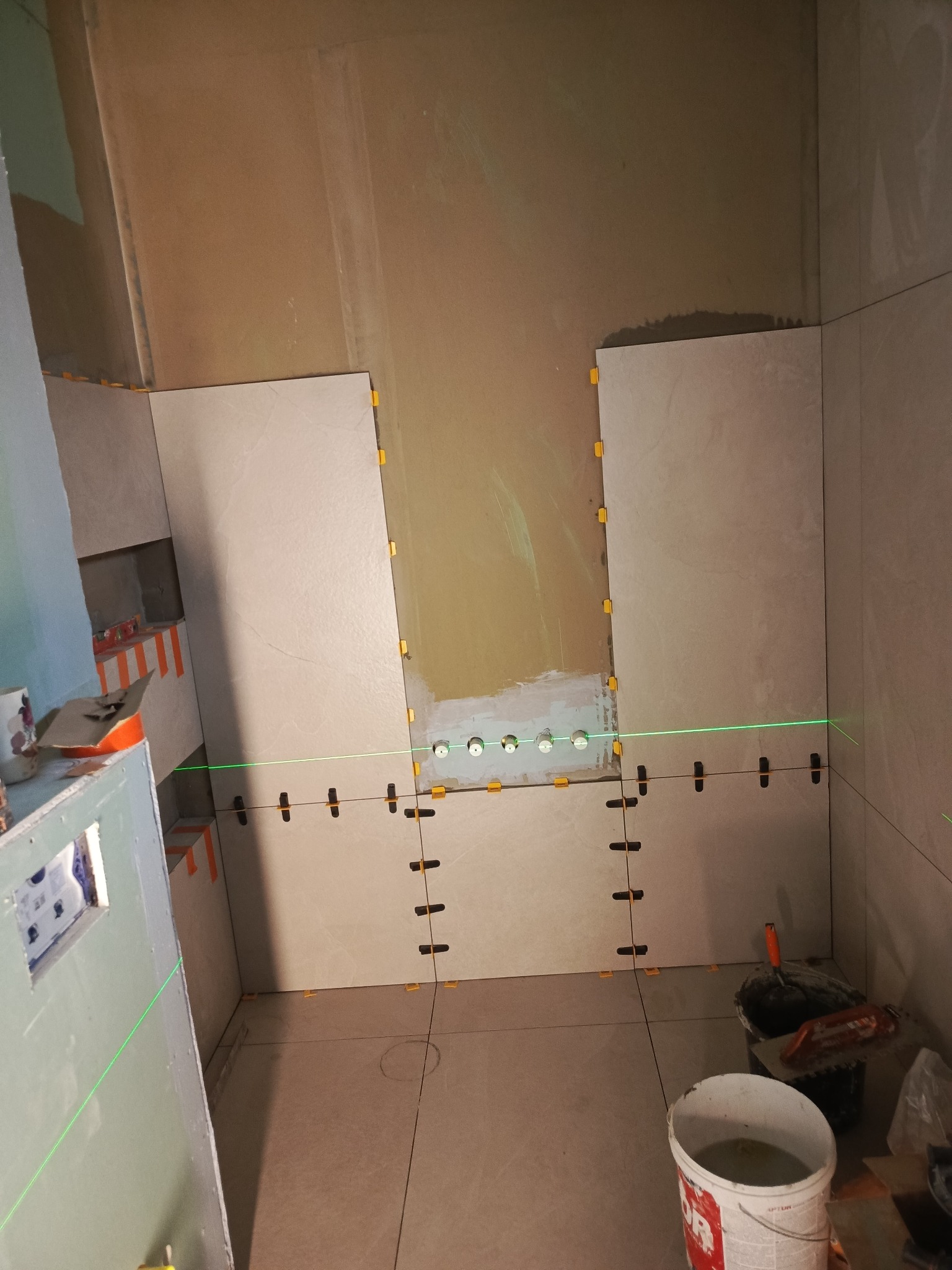 Układanie jasnych płytek w łazience, widoczny laser krzyżowy, poziomowanie płytek. Częściowo wykończone ściany, narzędzia glazurnicze w tle.