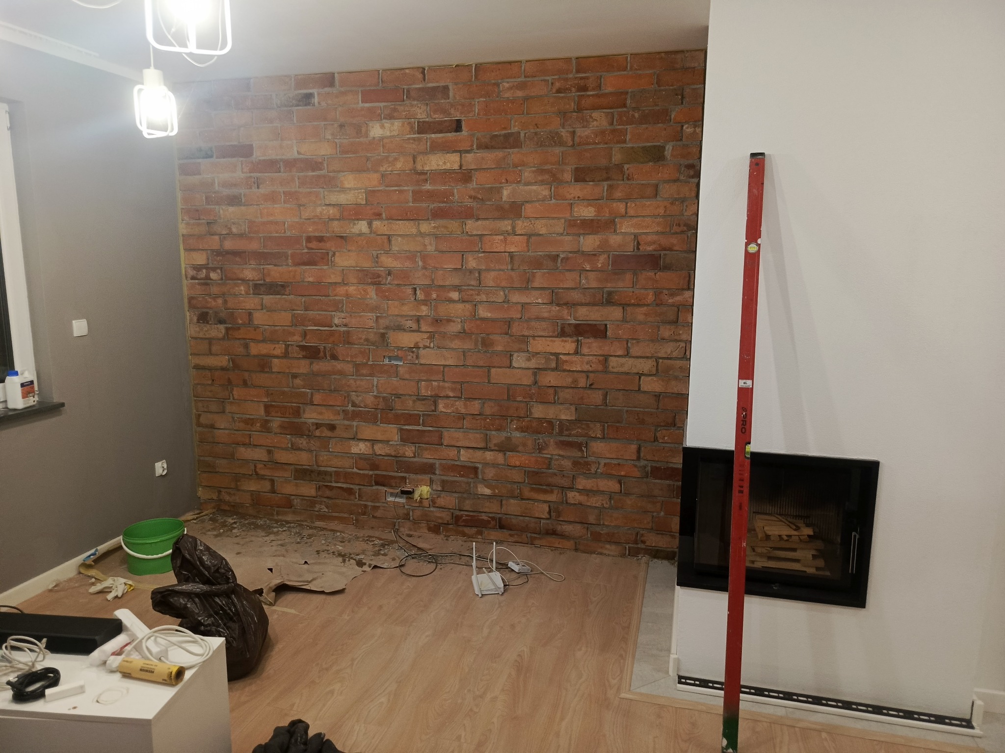Ściana z cegły w trakcie wykańczania, widoczny poziomica, podłoga z panelami, fragment kominka. W tle wiadro i worek na śmieci. Oświetlenie sufitowe.