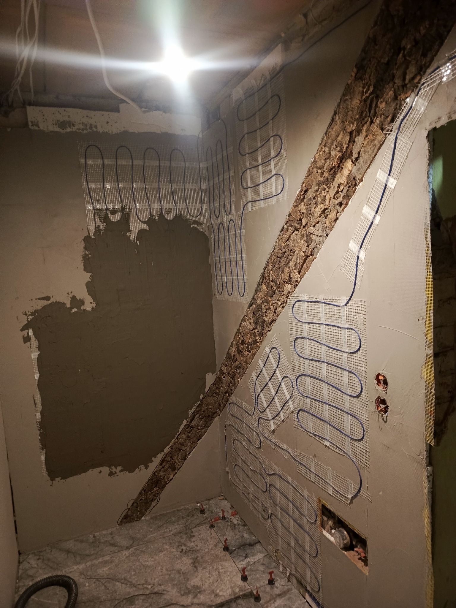 Montaż siatki grzewczej na ścianie w rogu pomieszczenia, przygotowanej do dalszych prac wykończeniowych. Widoczne kable i fragmenty instalacji elektrycznej.
