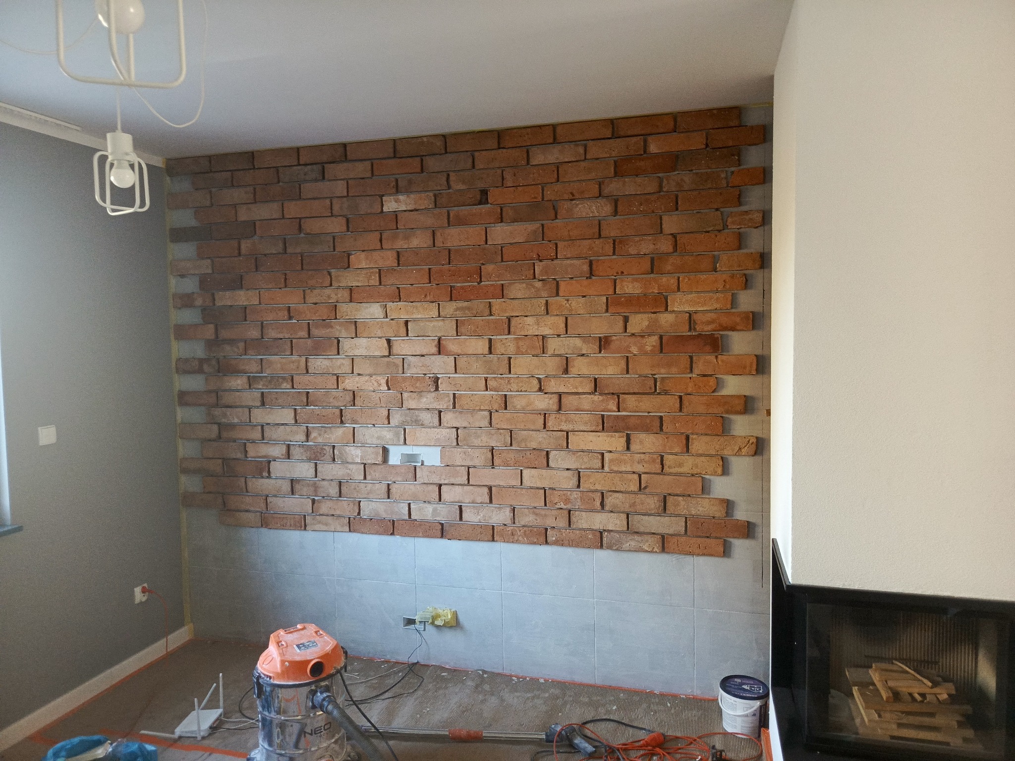 Ściana z cegły klinkierowej w trakcie montażu, widoczny fragment ściany z płytkami, odkurzacz i narzędzia. Częściowo wykończone wnętrze z lampą sufitową.