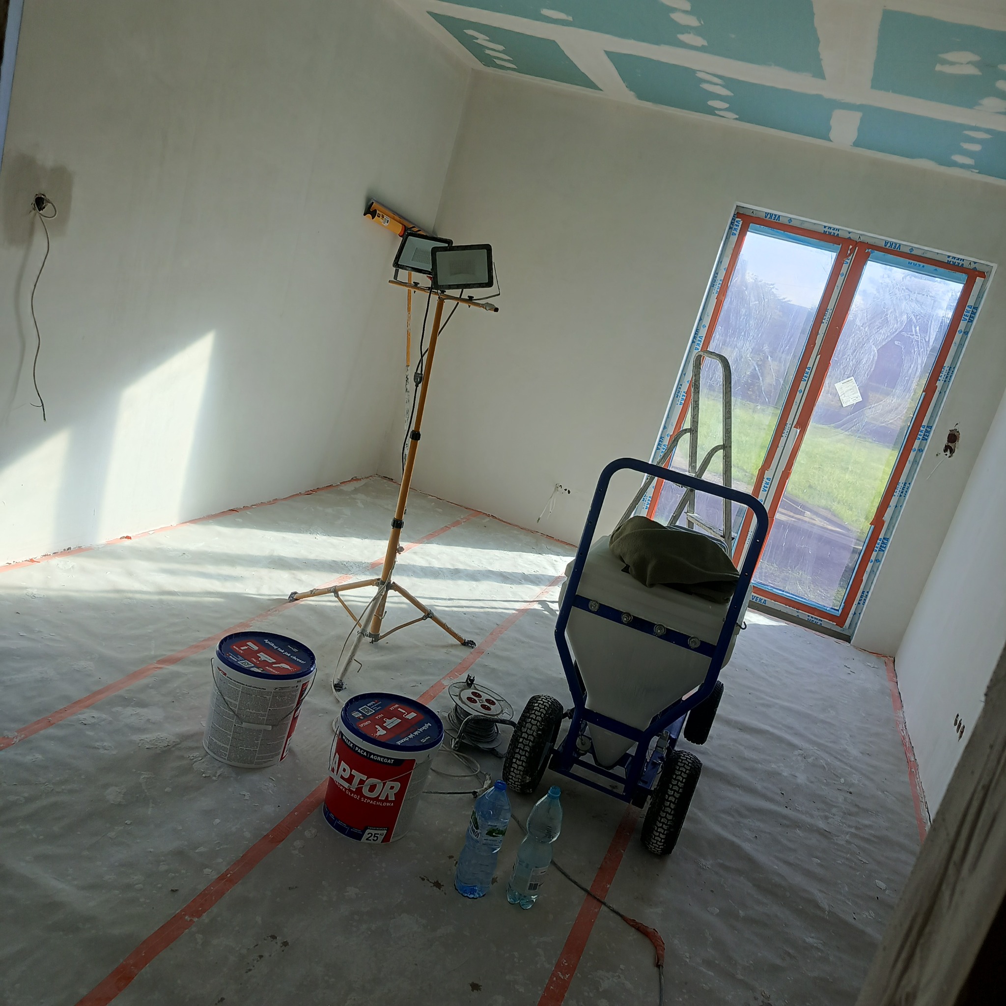 Wnętrze w trakcie malowania: ściany przygotowane, sufit z płyt karton-gips, agregat malarski, farby Raptor, lampa budowlana. Podłoga zabezpieczona folią.