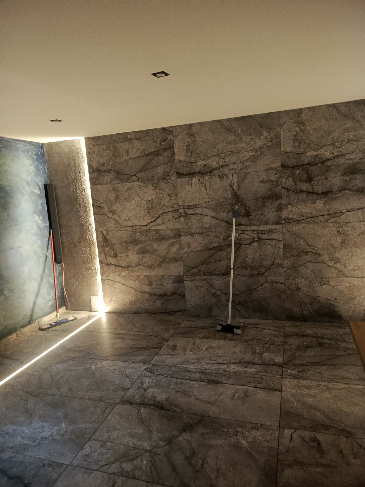 Wykończona łazienka z szarymi płytkami imitującymi marmur na ścianach i podłodze. Widoczna listwa LED wzdłuż ściany i mop. Minimalistyczny design.