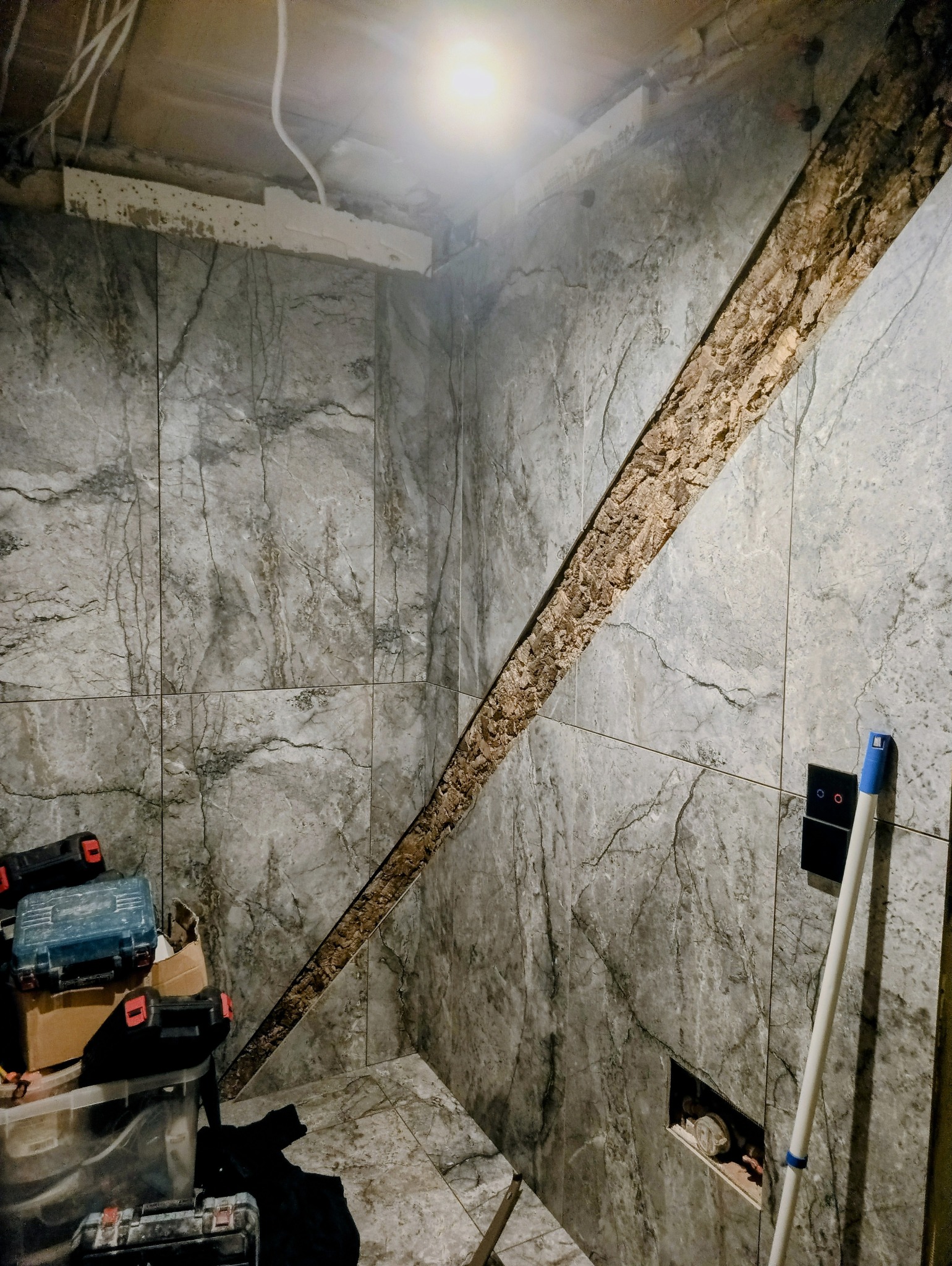 Ściana w trakcie wykańczania z szarymi płytkami imitującymi marmur, z widocznym fragmentem niezabezpieczonej ściany i narzędziami w rogu. Oświetlenie sufitowe.