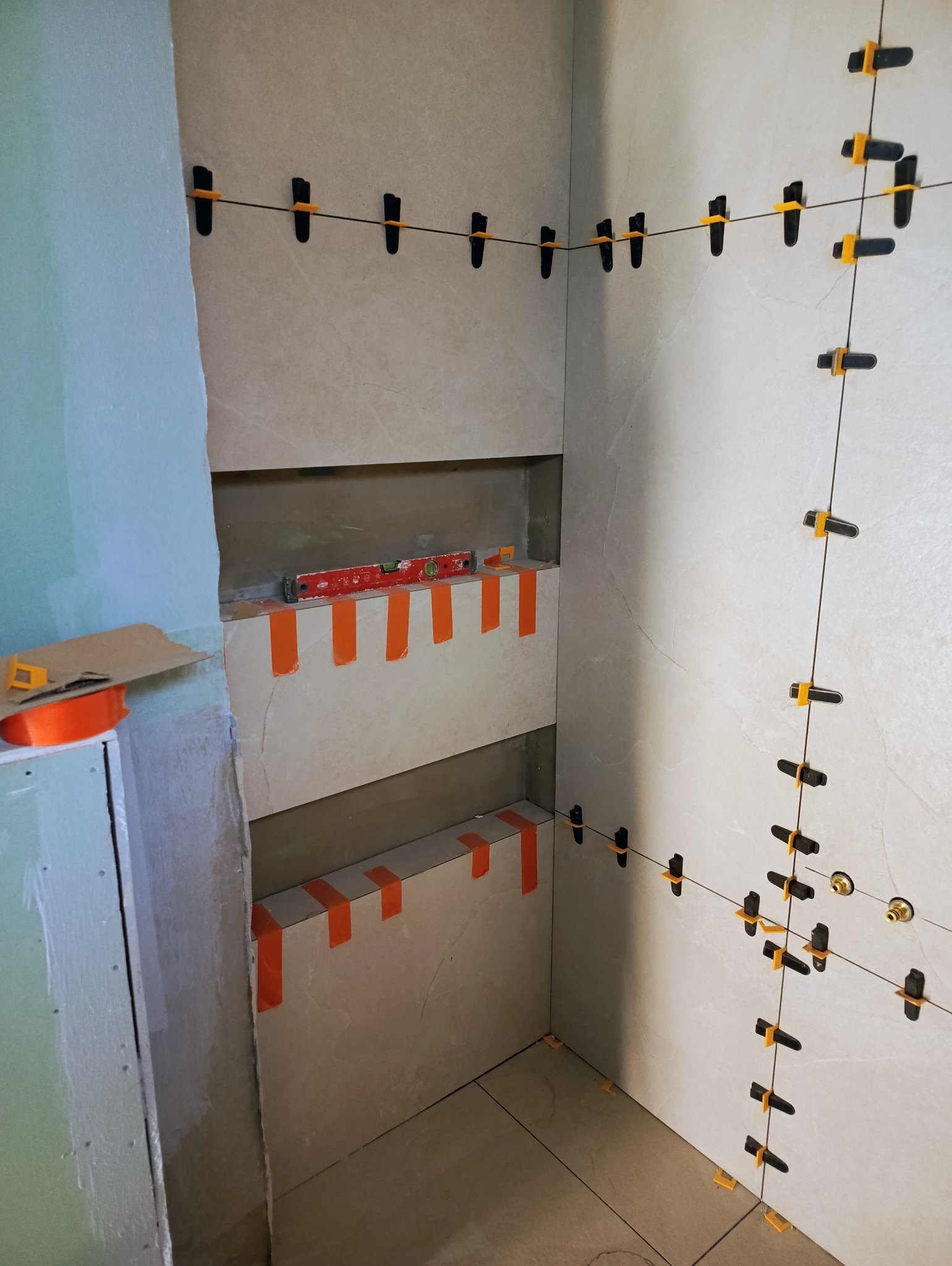 Układanie beżowych płytek w narożniku łazienki. Widoczne poziomice, taśmy i system poziomowania płytek. Wnęki w ścianie przygotowane pod półki.