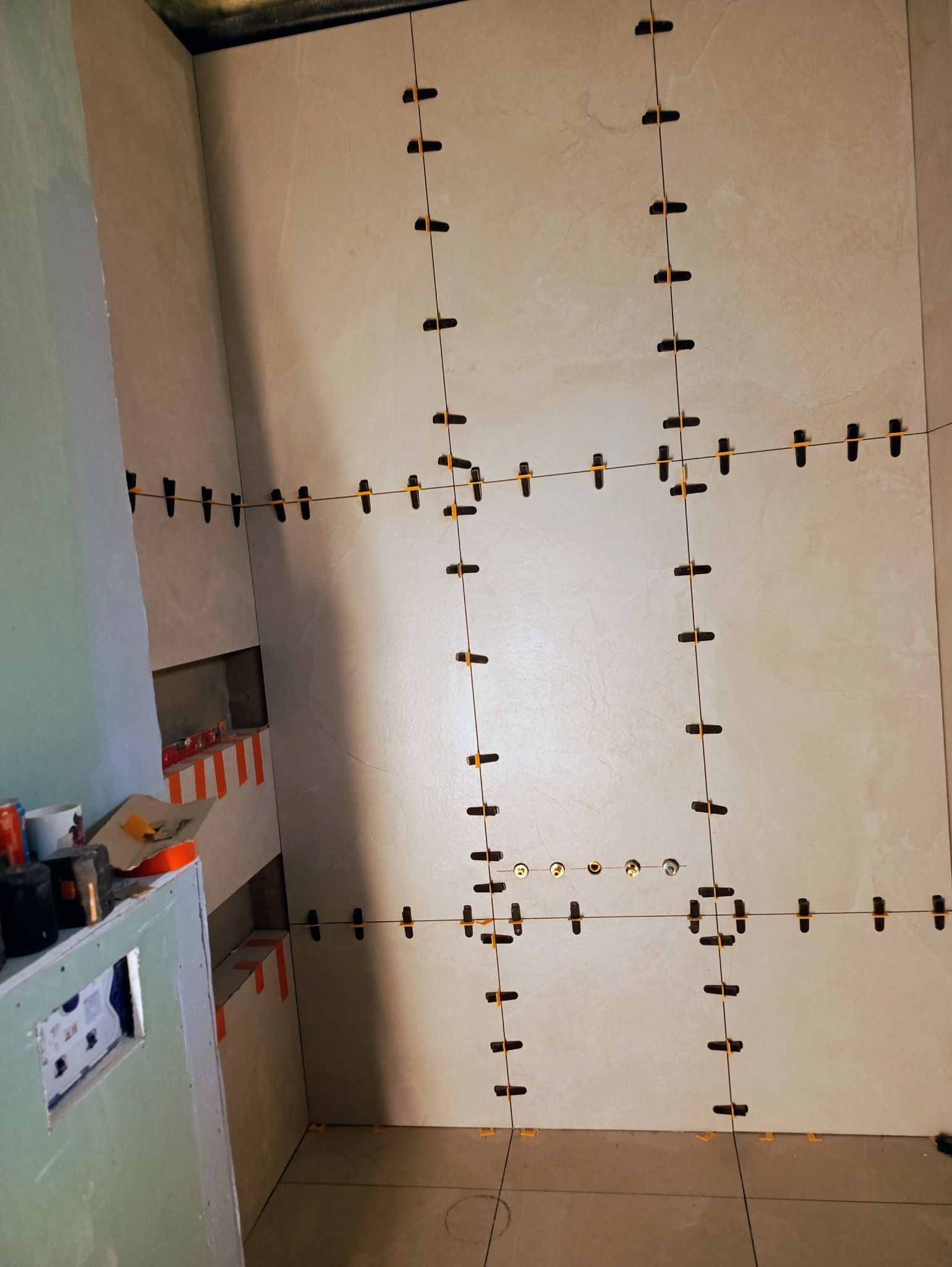 Ściana w trakcie układania płytek z użyciem systemu poziomowania. Widoczne krzyżyki dystansowe i fragment ściany z płytą karton-gips. Prace glazurnicze w łazience.