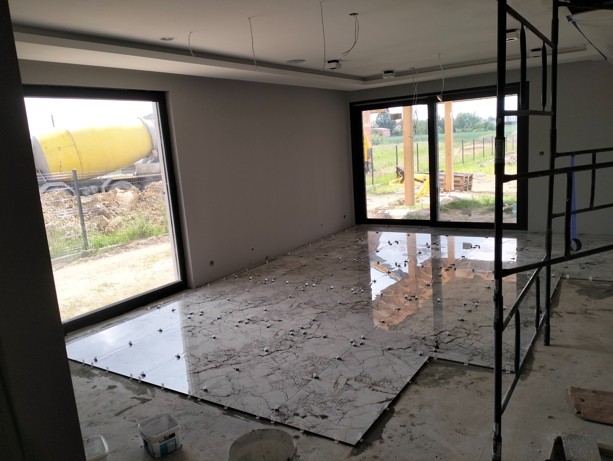 Układanie płytek marmurowych w jasnym wnętrzu z widokiem na betoniarkę za oknem. Widoczne krzyżyki dystansowe i rusztowanie.