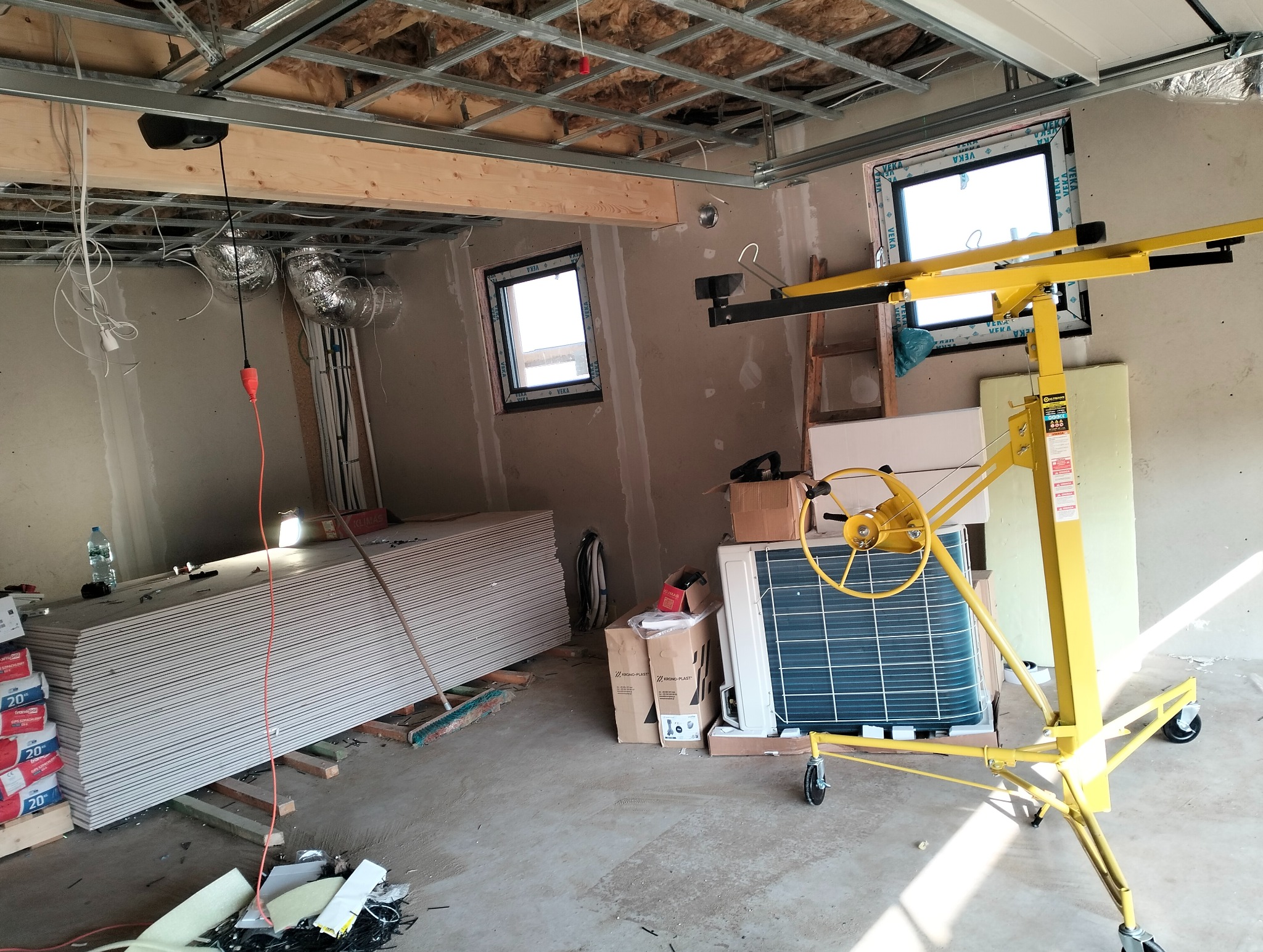 Wnętrze w trakcie prac z płytami gips-karton, widoczny podnośnik do płyt, profile sufitowe i okna. Na podłodze ułożone płyty g-k i materiały budowlane.