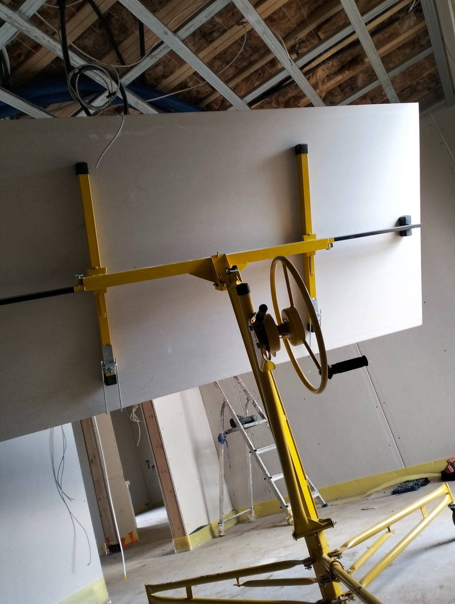 Żółty podnośnik do płyt KG podtrzymuje białą płytę na tle surowego wnętrza z widoczną konstrukcją sufitu i ścianami w trakcie montażu płyt kartonowo-gipsowych.