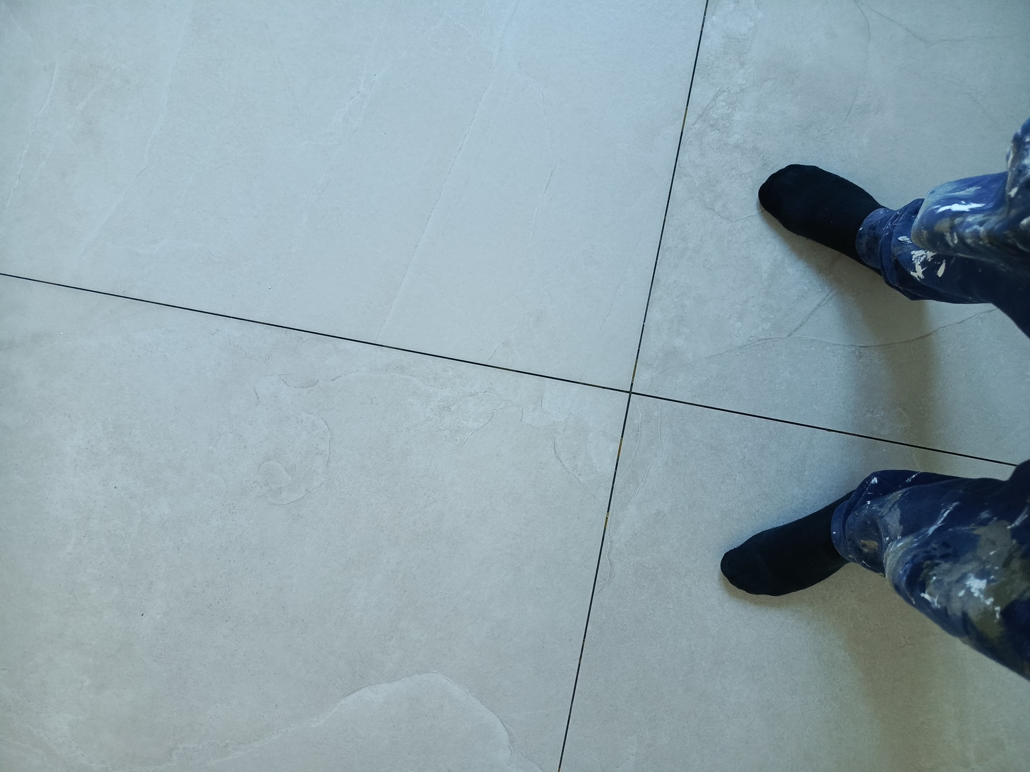 Jasnoszare płytki podłogowe z fugami, widoczne stopy w czarnych skarpetach i spodnie robocze z plamami farby. Perspektywa z góry.