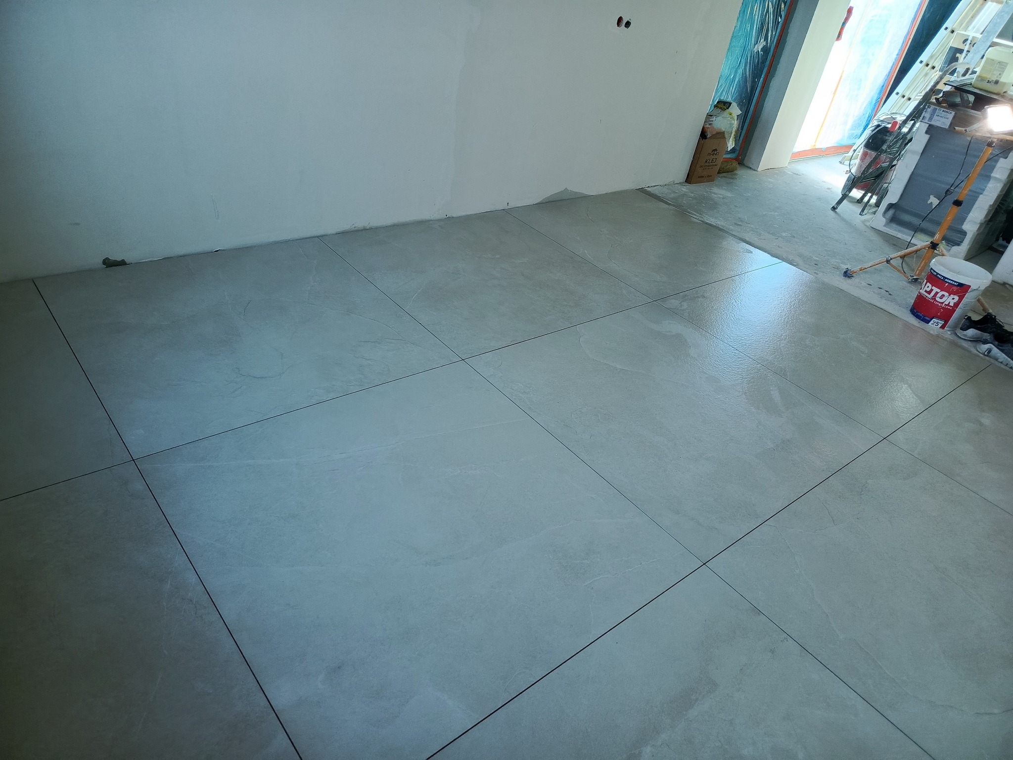 Ułożone, jasnoszare płytki podłogowe w nowym wnętrzu. Widoczne narzędzia i materiały budowlane w tle. Świeżo położona podłoga z płytek.