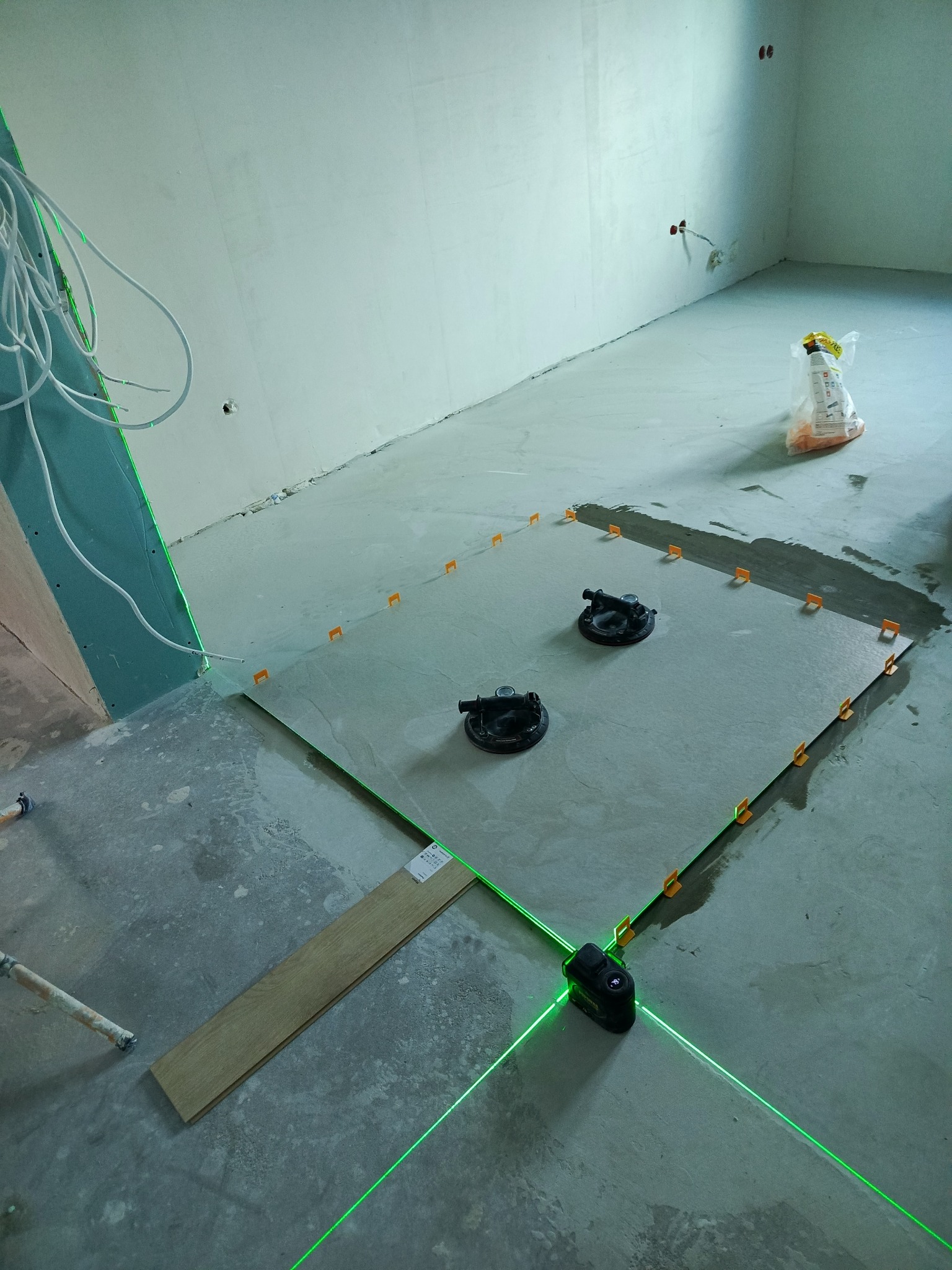 Układanie płytek podłogowych z użyciem lasera krzyżowego i przyssawek. Widoczne płytki, krzyżyki dystansowe i worek z zaprawą. Ściany surowe, w trakcie prac wykończeniowych.