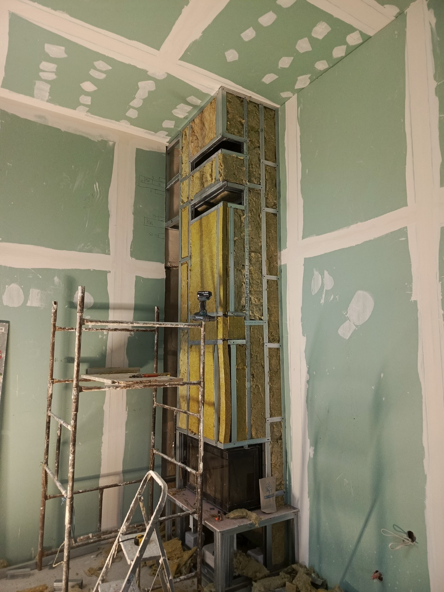 Montaż obudowy kominka z płyt gipsowo-kartonowych na metalowym stelażu, widoczna izolacja z wełny mineralnej, szpachlowane ściany i sufit, rusztowanie.