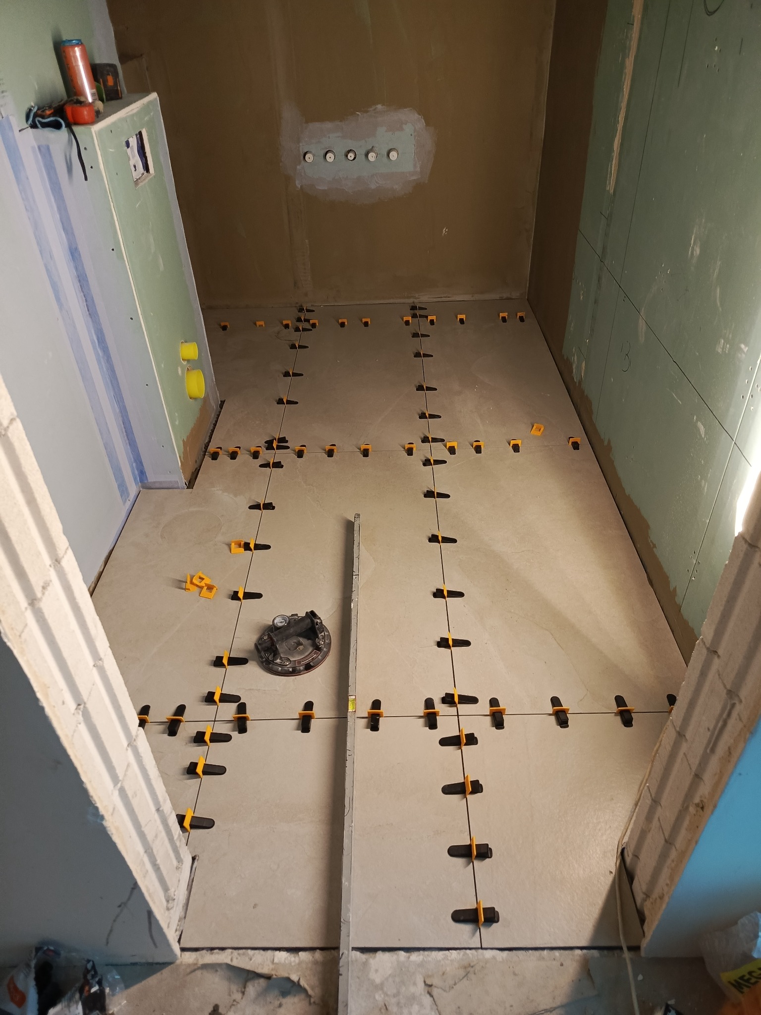 Układanie płytek podłogowych w łazience z użyciem systemu poziomowania. Widoczny przyrząd do przyssawek i poziomica. Ściany przygotowane pod wykończenie.