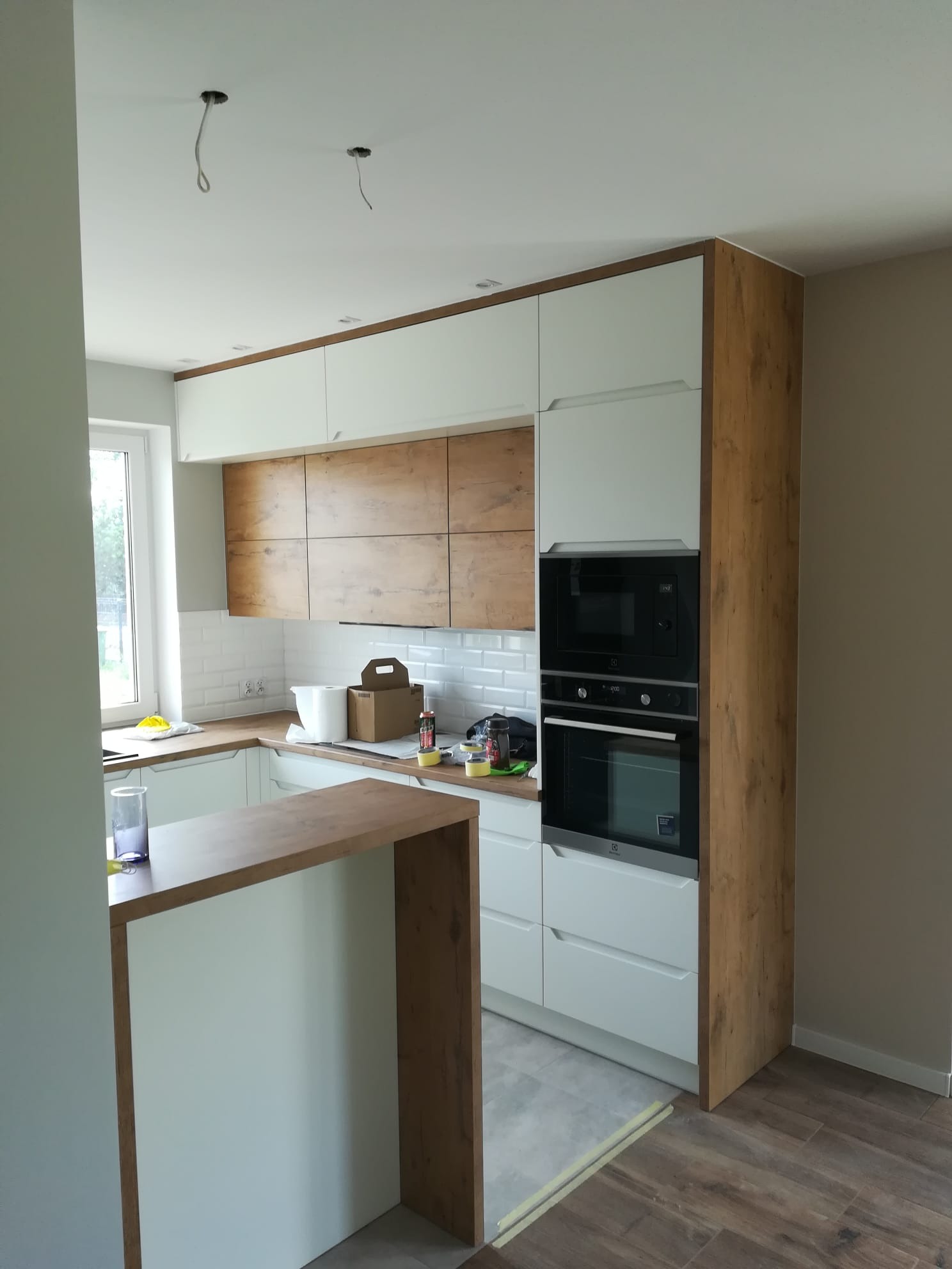Jasna kuchnia z drewnianymi akcentami: białe szafki, drewniane fronty górne i obudowa piekarnika. Widoczne niedokończone elementy wykończenia.