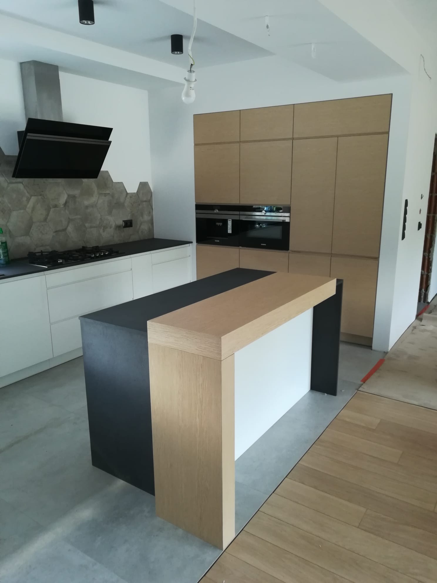 Nowoczesna kuchnia z wyspą, jasne szafki, heksagonalne płytki nad blatem, czarny okap i płyta. Podłoga częściowo wykończona panelami.