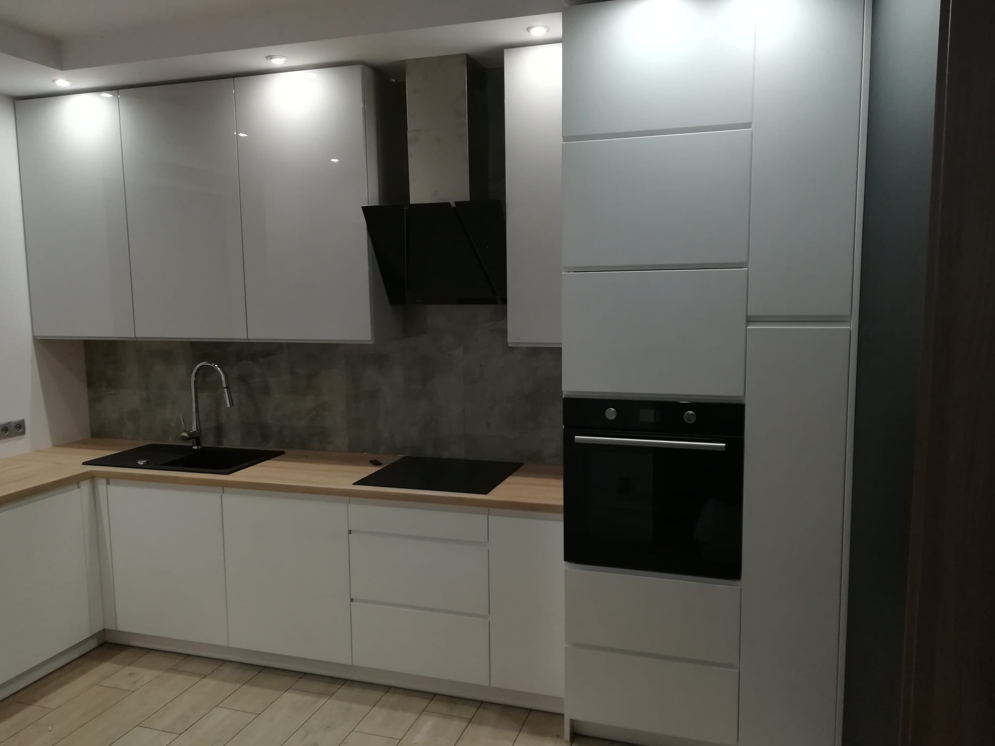 Nowoczesna kuchnia z białymi, błyszczącymi szafkami, drewnianym blatem i czarnym zlewem. Widoczny piekarnik i okap. Minimalistyczny design, szare płytki na ścianie.