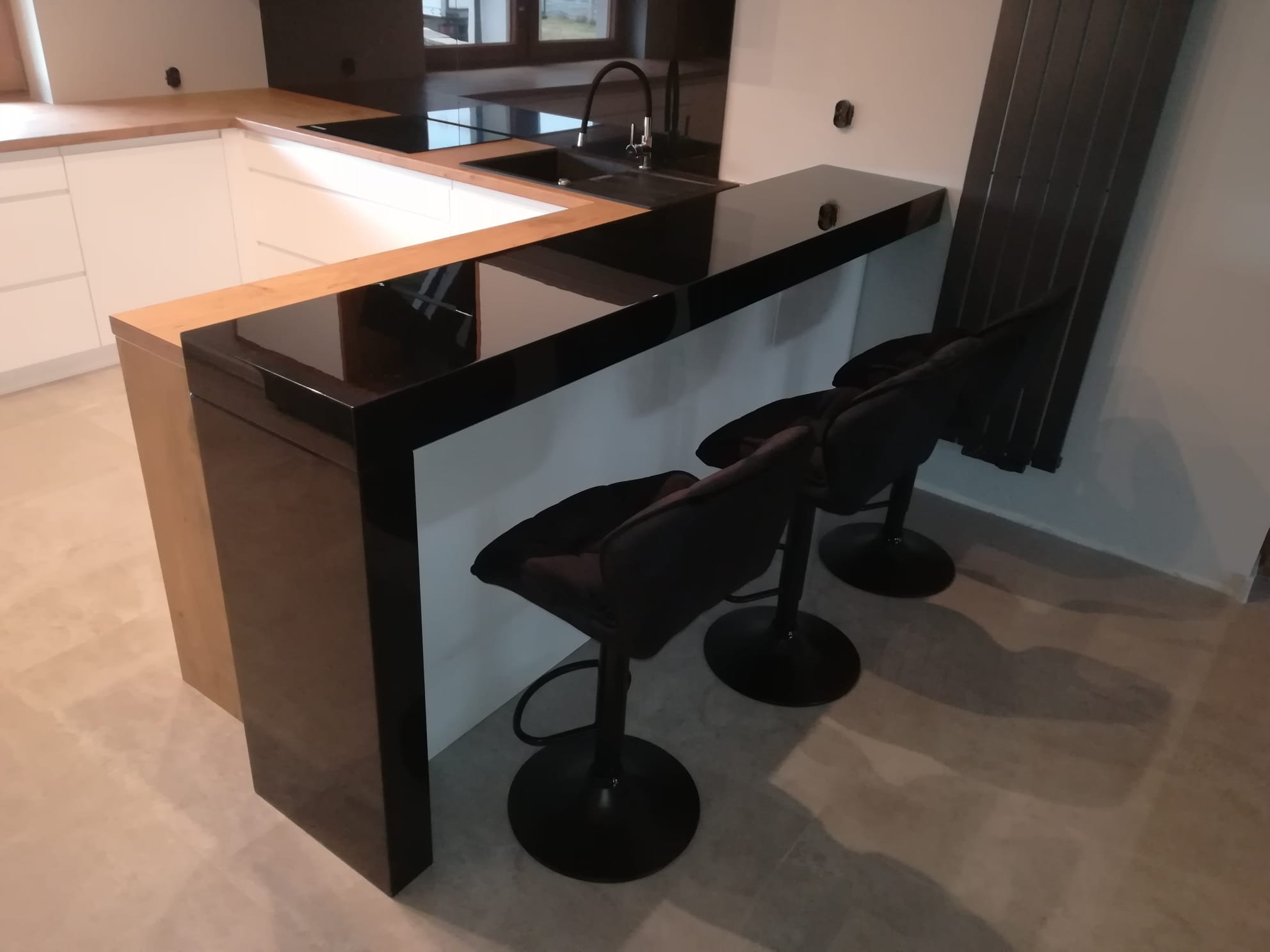 Nowoczesna kuchnia z czarnym blatem i barkiem, uzupełniona o trzy czarne hokery. Widoczne białe szafki i drewniane elementy wykończeniowe.