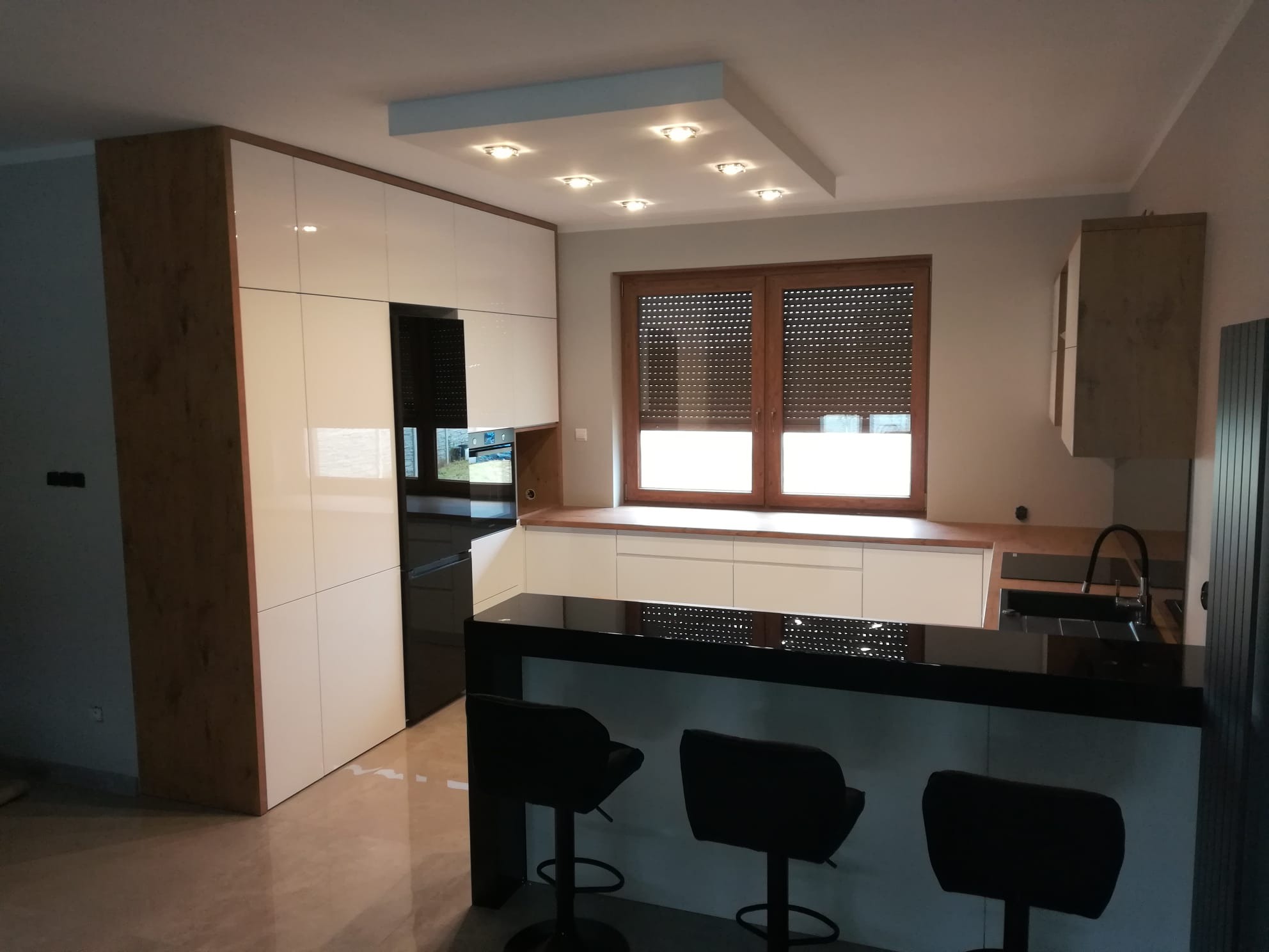 Nowoczesna kuchnia z białymi szafkami, czarnym blatem i drewnianymi akcentami. Widoczne trzy czarne hokery przy blacie. Okno z drewnianą ramą i roletami.