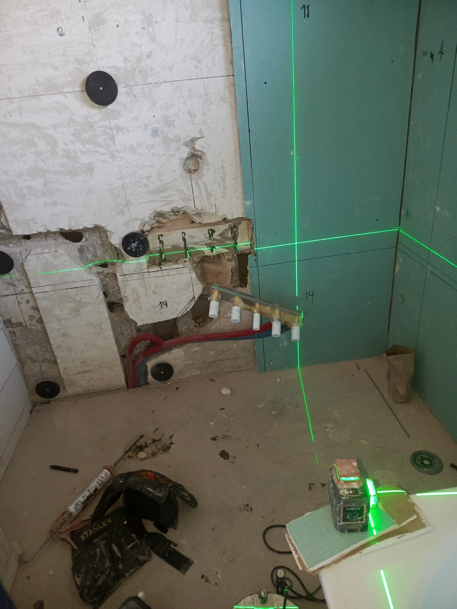 Nierówna ściana z fragmentami płyty gipsowej, widoczne rury i laser krzyżowy wyznaczający poziom. Na podłodze narzędzia i resztki materiałów budowlanych.