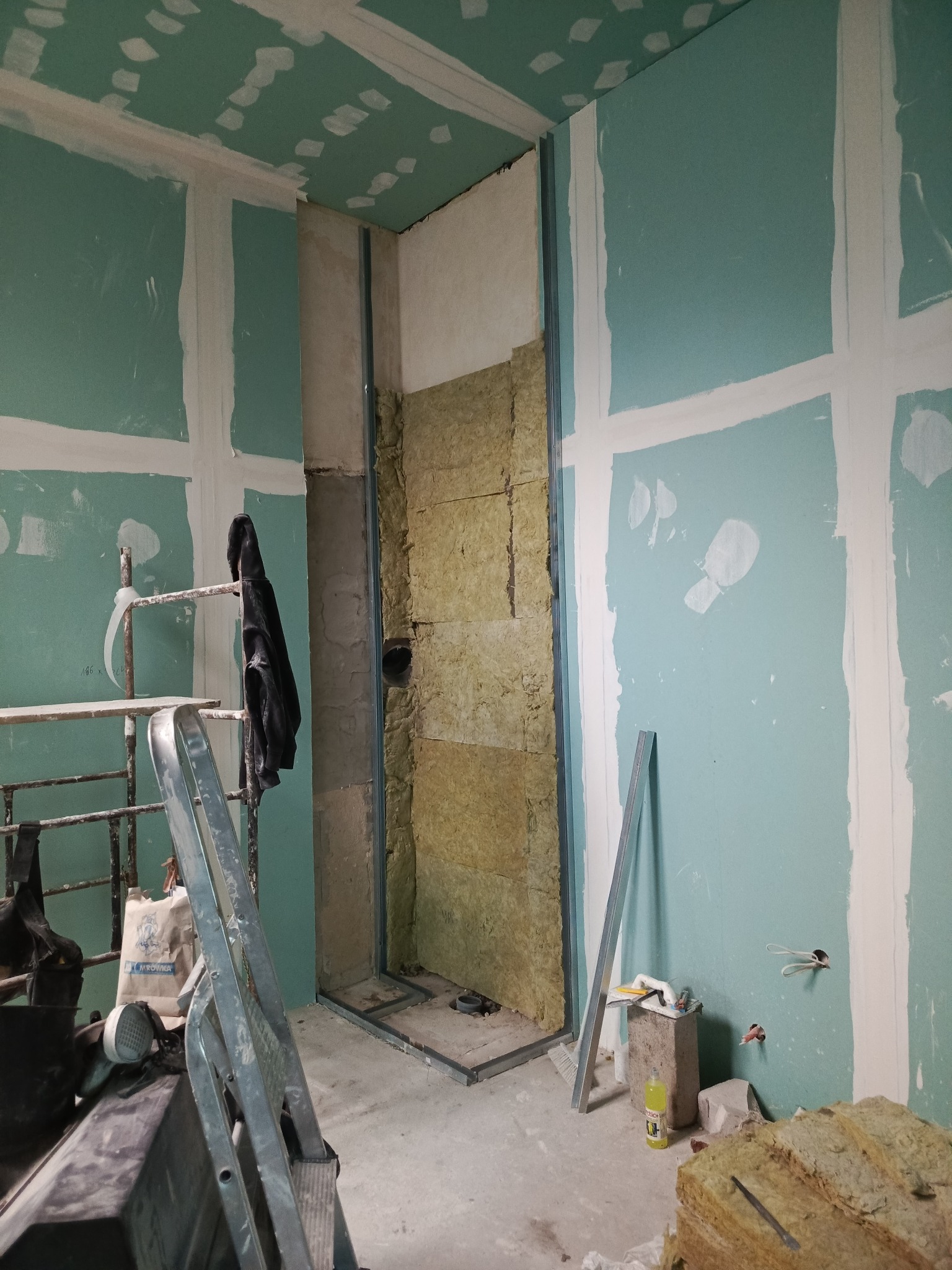 Ściana z płyt gipsowo-kartonowych w trakcie montażu, widoczne profile i izolacja. Szpachlowane łączenia płyt. W tle rusztowanie i narzędzia.