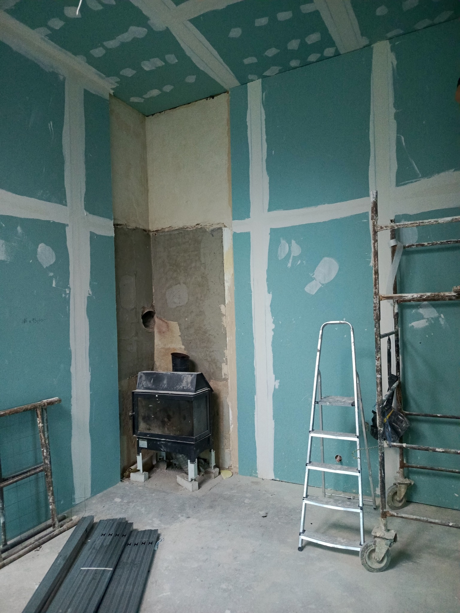 Pokój w trakcie wykańczania: ściany z płyt karton-gips, widoczne łączenia, piec wolnostojący, drabina i rusztowanie. Surowy stan, przygotowanie do dalszych prac.