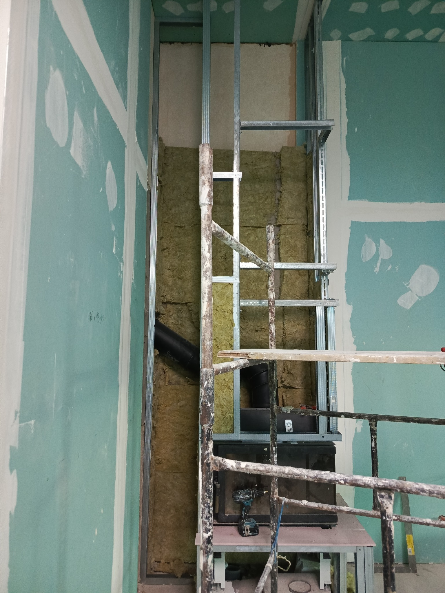 Ściana z izolacją z wełny mineralnej, metalowym stelażem i częściowo zamontowaną płytą gipsowo-kartonową. Widoczne narzędzia na rusztowaniu.