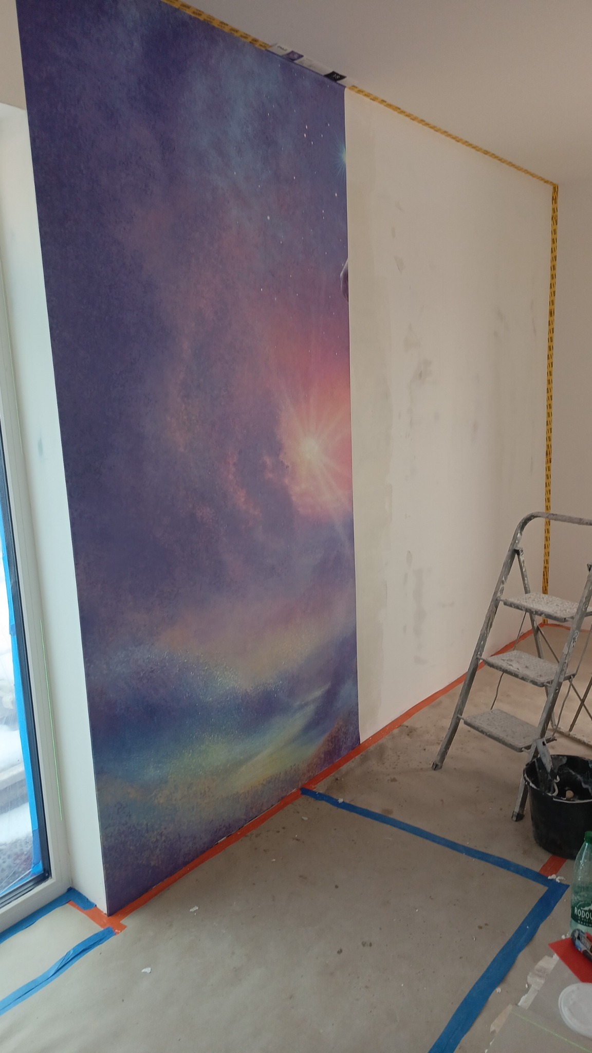 Montaż fototapety z motywem kosmosu na ścianie, widoczne narzędzia i drabina, zabezpieczona podłoga taśmą malarską, etap prac wykończeniowych.