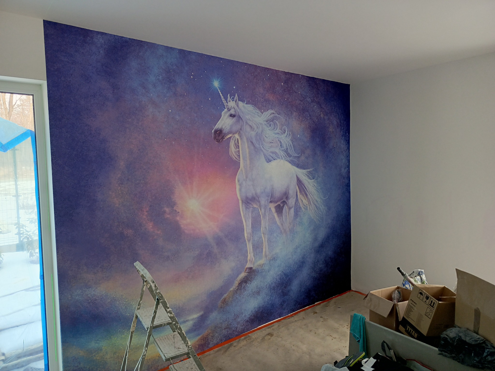 Ściana z fototapetą przedstawiającą jednorożca na tle kosmosu, drabina malarska i kartony na podłodze. Wykończenie wnętrza w trakcie realizacji.