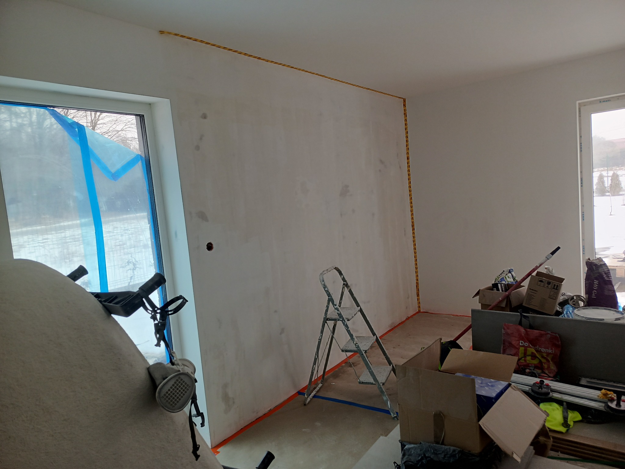 Przygotowania do malowania ścian w jasnym wnętrzu. Oklejone okno, drabina, taśmy malarskie i narzędzia malarskie. Widoczne ślady po szpachlowaniu na ścianie.