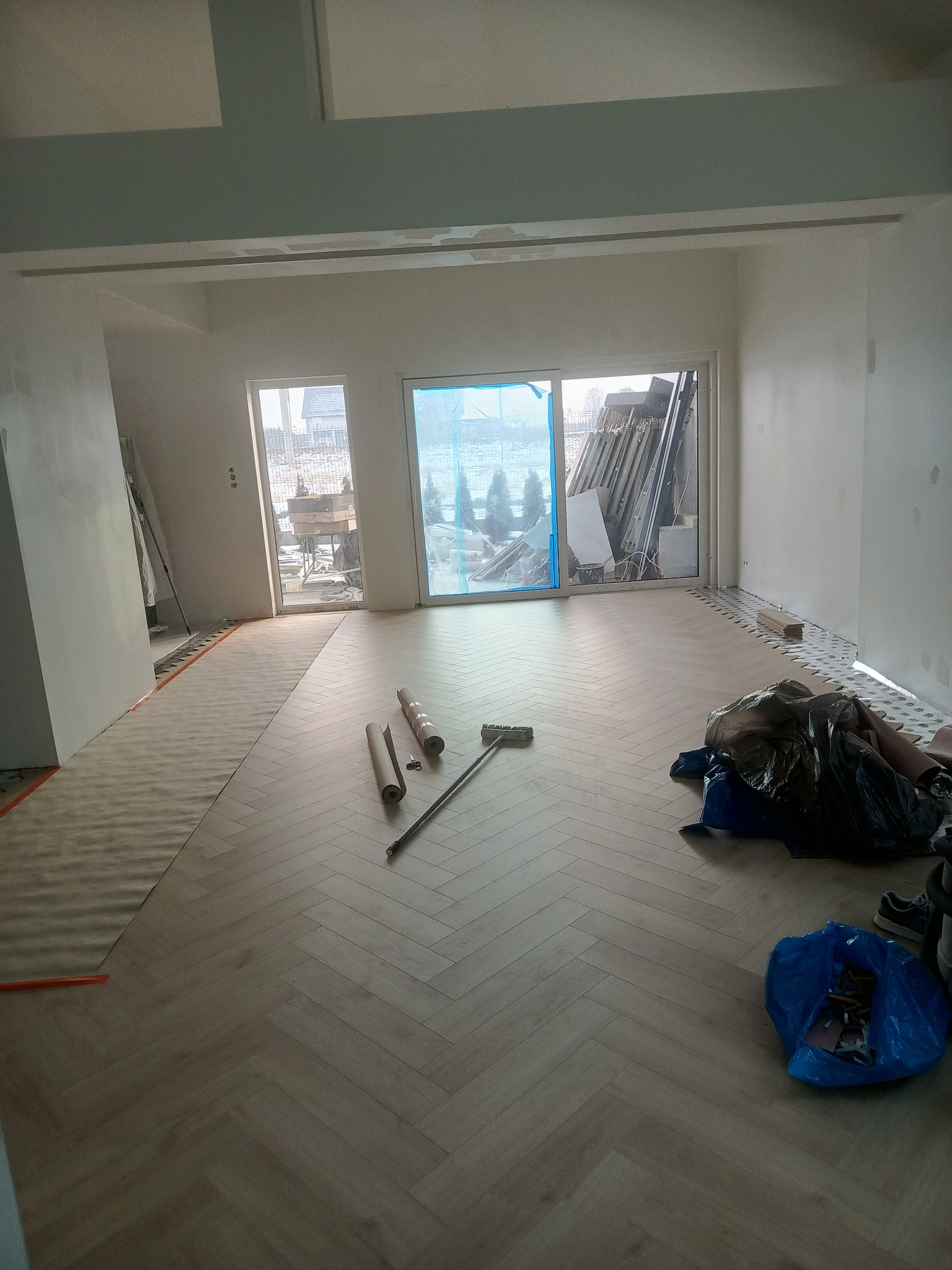 Jasny parkiet ułożony w jodełkę w przestronnym, jasnym pokoju w trakcie wykańczania. Widoczne materiały budowlane i narzędzia.