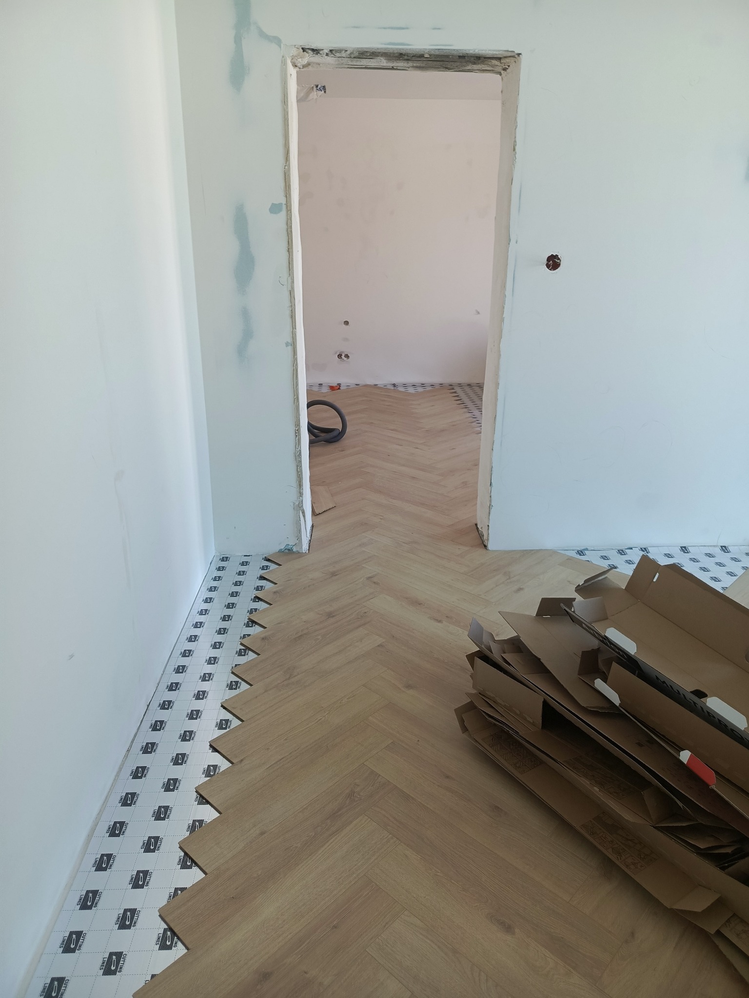Układanie paneli podłogowych w jodełkę, widoczne podkłady i kartony po panelach. Ściany przygotowane do malowania, surowy otwór drzwiowy.