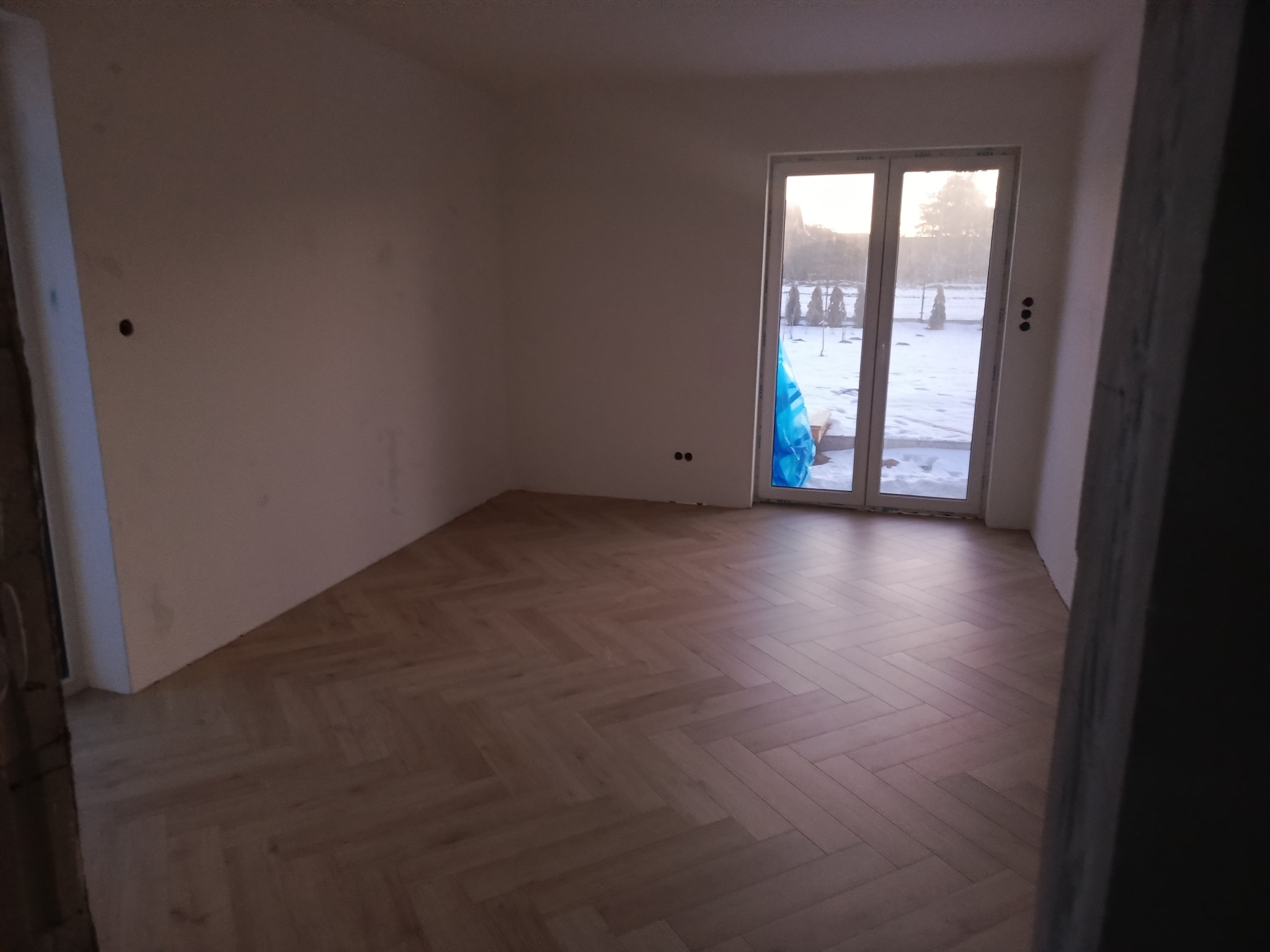 Jasne, parkietowe panele ułożone w jodełkę w pustym pokoju z oknem na zimowy krajobraz. Ściany przygotowane do malowania, widoczne punkty elektryczne.