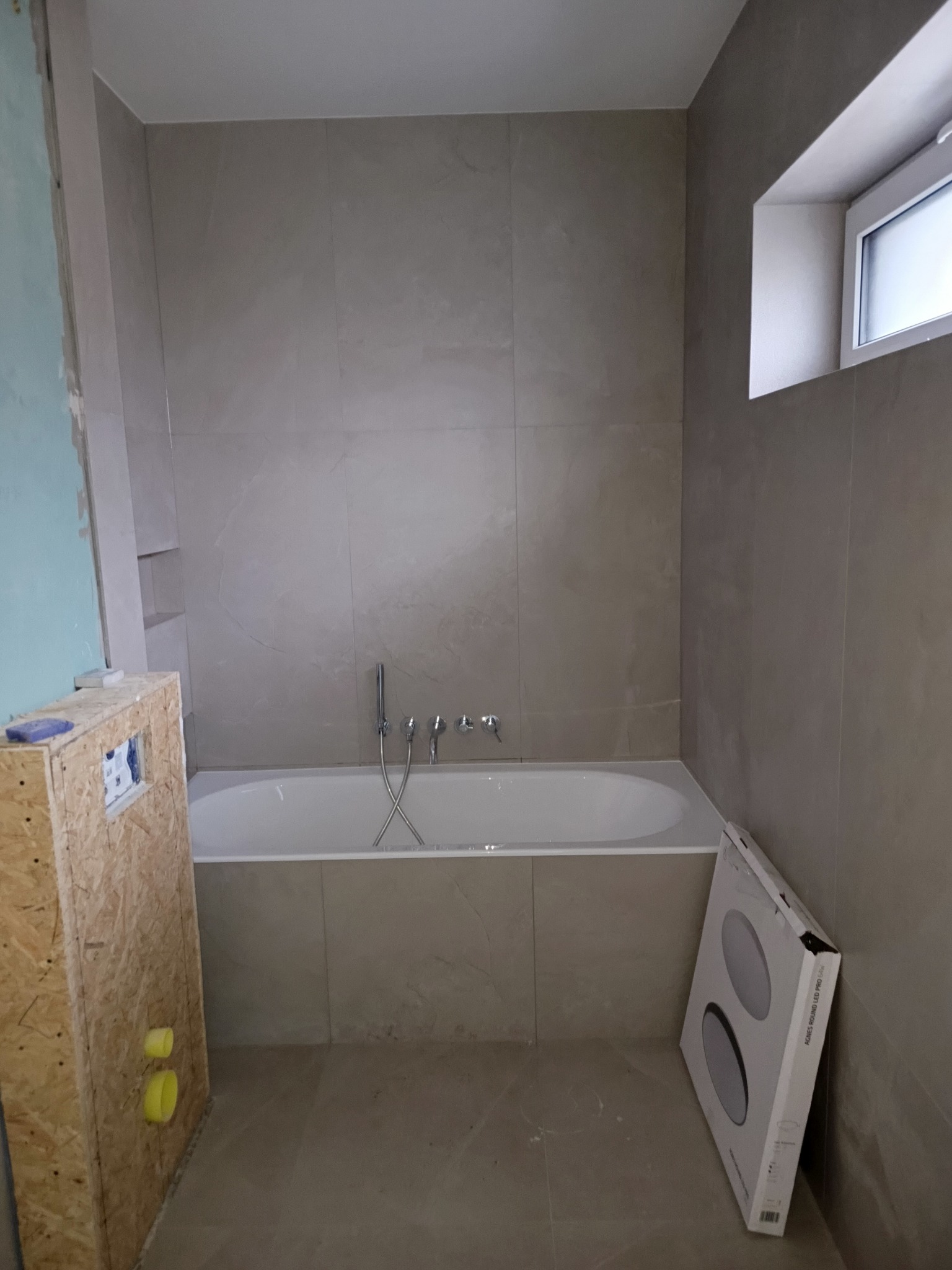 Wykończona łazienka z wanną w zabudowie, płytki w odcieniach beżu, okno. Częściowo widoczna obudowa instalacji i karton z lampami. Ujęcie z góry, neutralne oświetlenie.