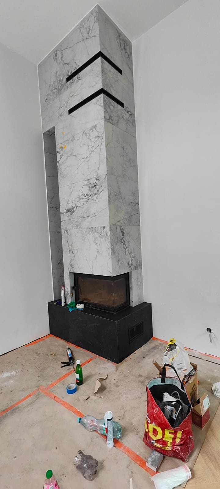 Obudowa kominka z marmurowymi płytami w trakcie montażu. Widoczne czarne, poziome akcenty dekoracyjne i narzędzia na podłodze. Pomieszczenie w stanie surowym.