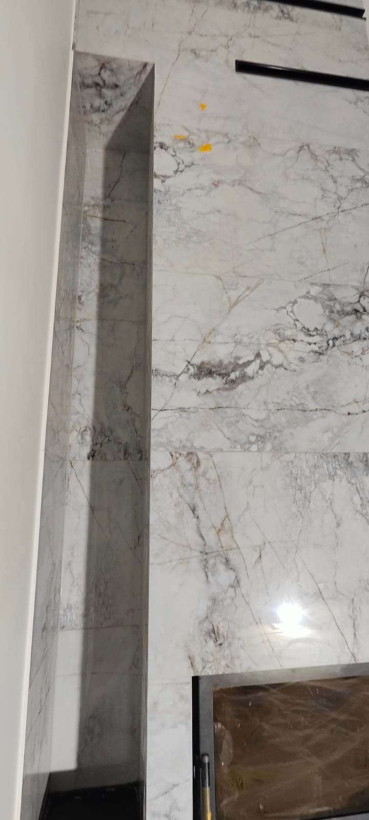 Ściana wyłożona białymi płytkami imitującymi marmur z wnęką i czarnymi elementami dekoracyjnymi. Widoczne żółte taśmy zabezpieczające.