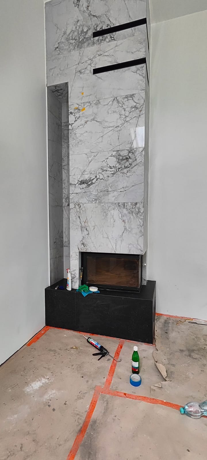 Obudowa kominka z płyt marmurowych z czarnymi akcentami, widok z dołu, na podłodze narzędzia i materiały budowlane, pomarańczowe linie wyznaczają obszar roboczy.