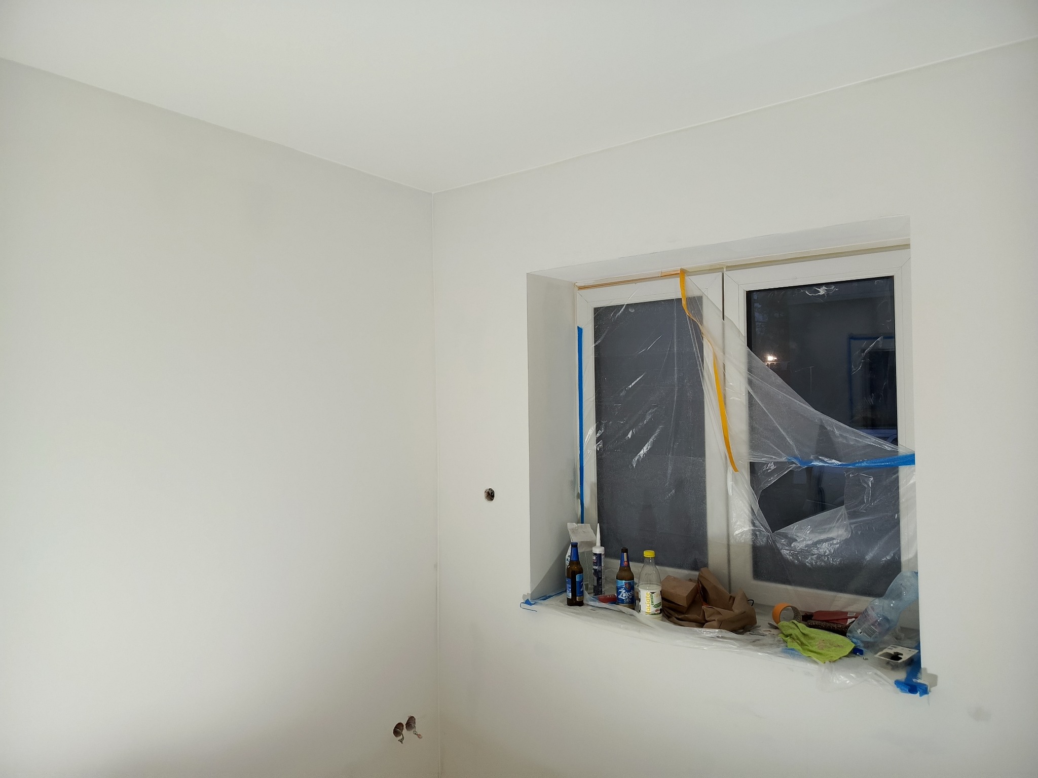Białe ściany po malowaniu, okno zabezpieczone folią malarską, widoczne ślady prac malarskich i narzędzia na parapecie. Minimalistyczne wnętrze po odświeżeniu.